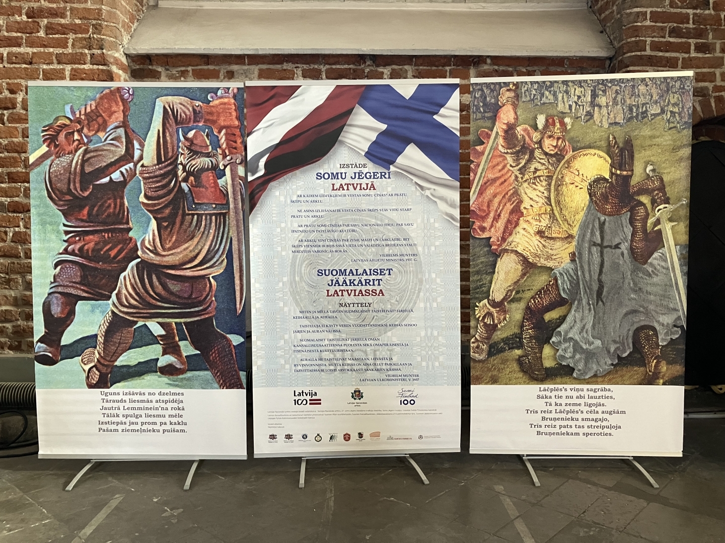 Krāsaina bilde, izstādes atklāšanas stendi, pa kreisi ilustrācija no Somu eposa Kelevala, pa labi - no Lāčplēša