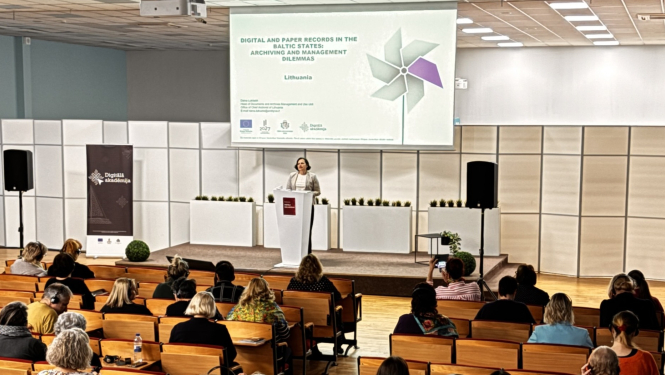 Konferenču zāle, foto centrā pie pults sieviete-lektore, fonā projecēts referāta nosaukums. Zālē klausītāji, visi ar skatu pret lektori. 