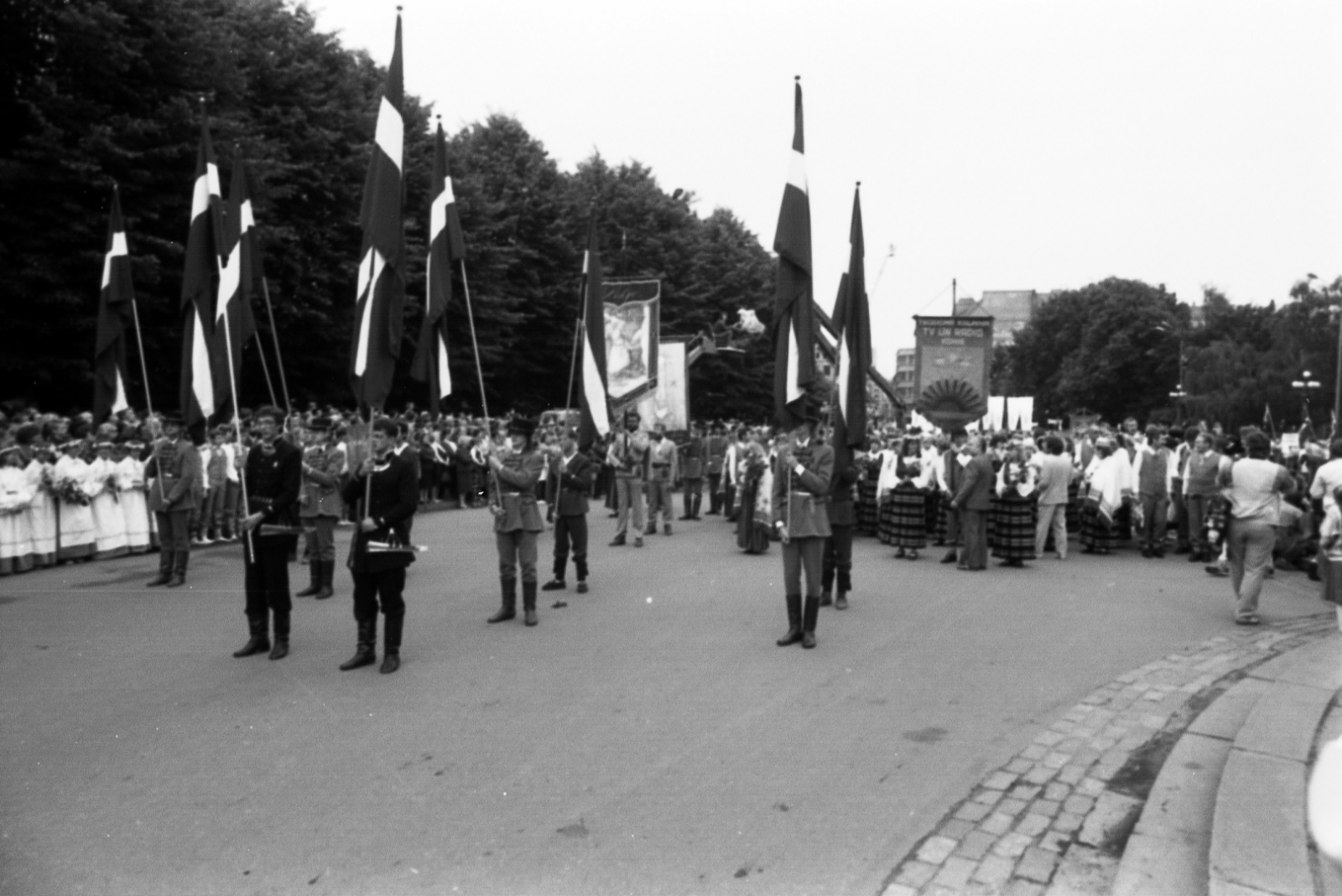Melnbalta bilde no izstādes "Dziesmu spēks" - kadrs no gājiena pie Brīvības pieminekļa, karogi un Dziesmu svētku karogs, kolonnas sākums