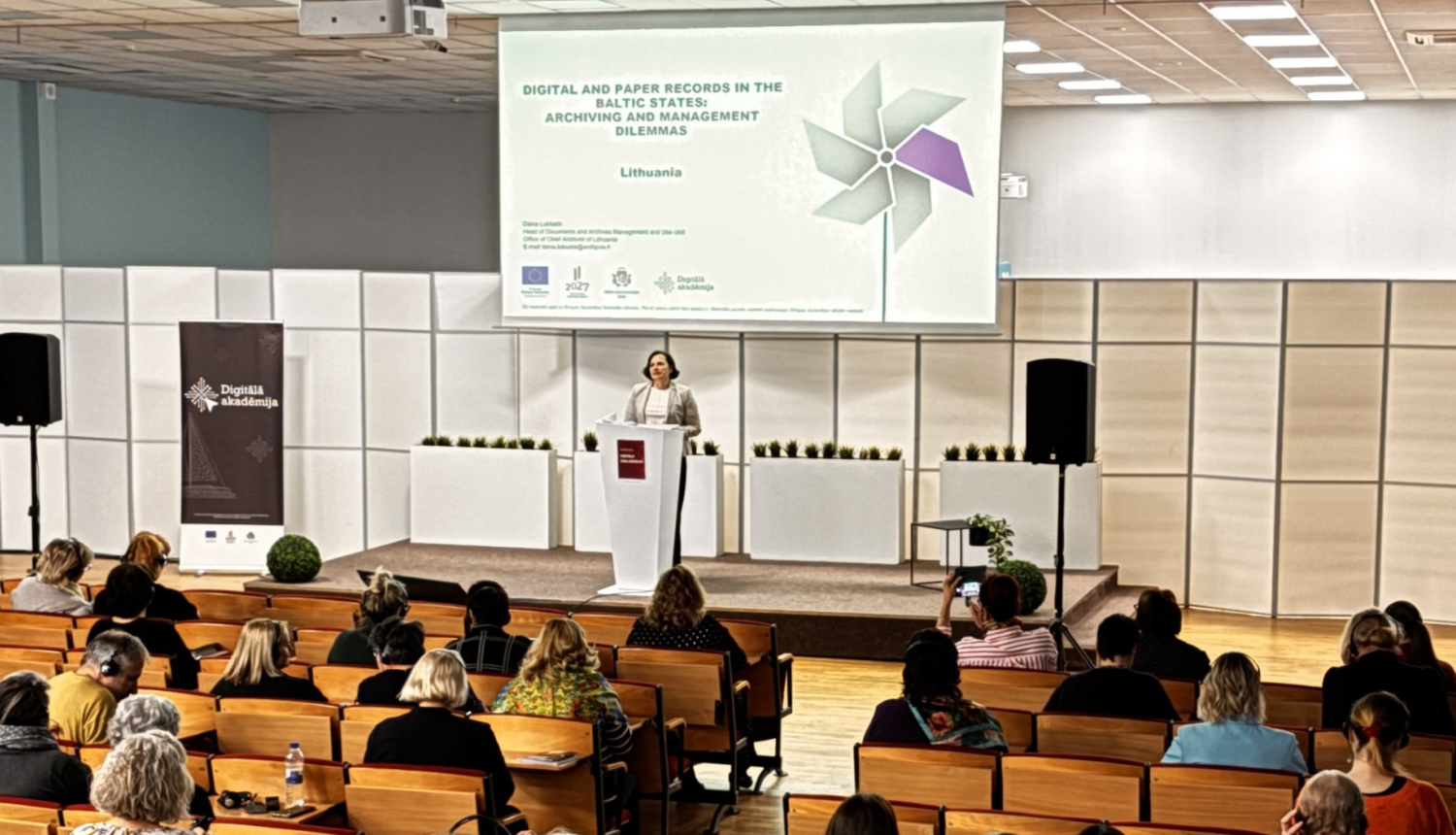 Konferenču zāle, foto centrā pie pults sieviete-lektore, fonā projecēts referāta nosaukums. Zālē klausītāji, visi ar skatu pret lektori. 