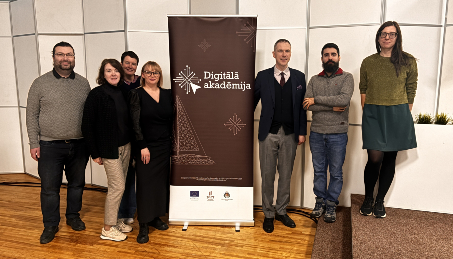 Grupas foto, no kreisās puses stāv četri cilvēki, pa vidu plakāts "Digitālā akadēmija", pa labi trīs cilvēki 