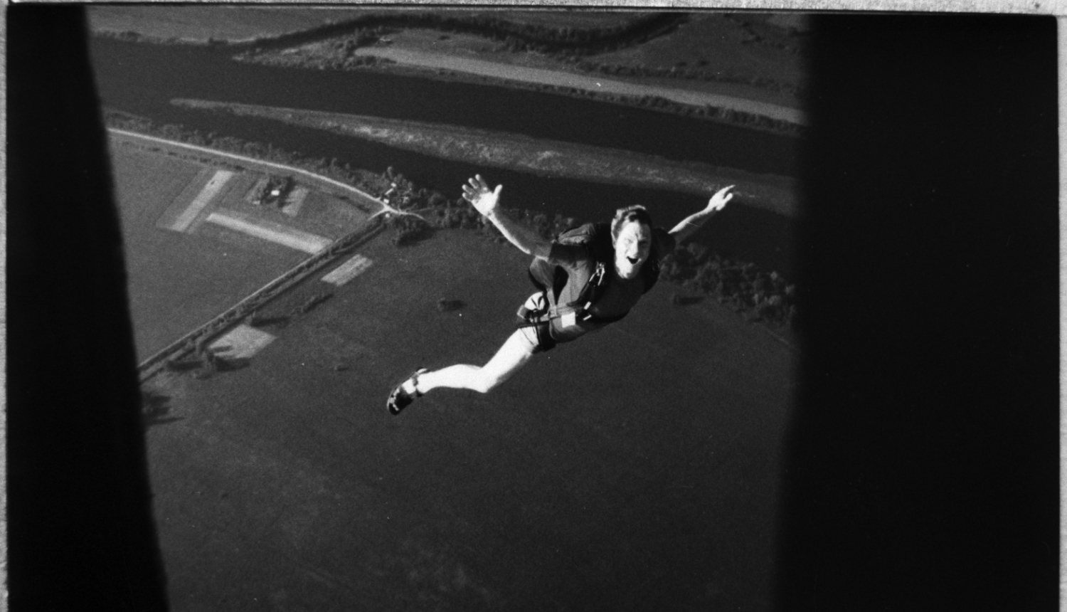 Melnbalta bilde, cilvēks lēcienā no lidmašīnas, smaida, atplestām rokām, virs laukiem, izpletnis vēl nav atvērts