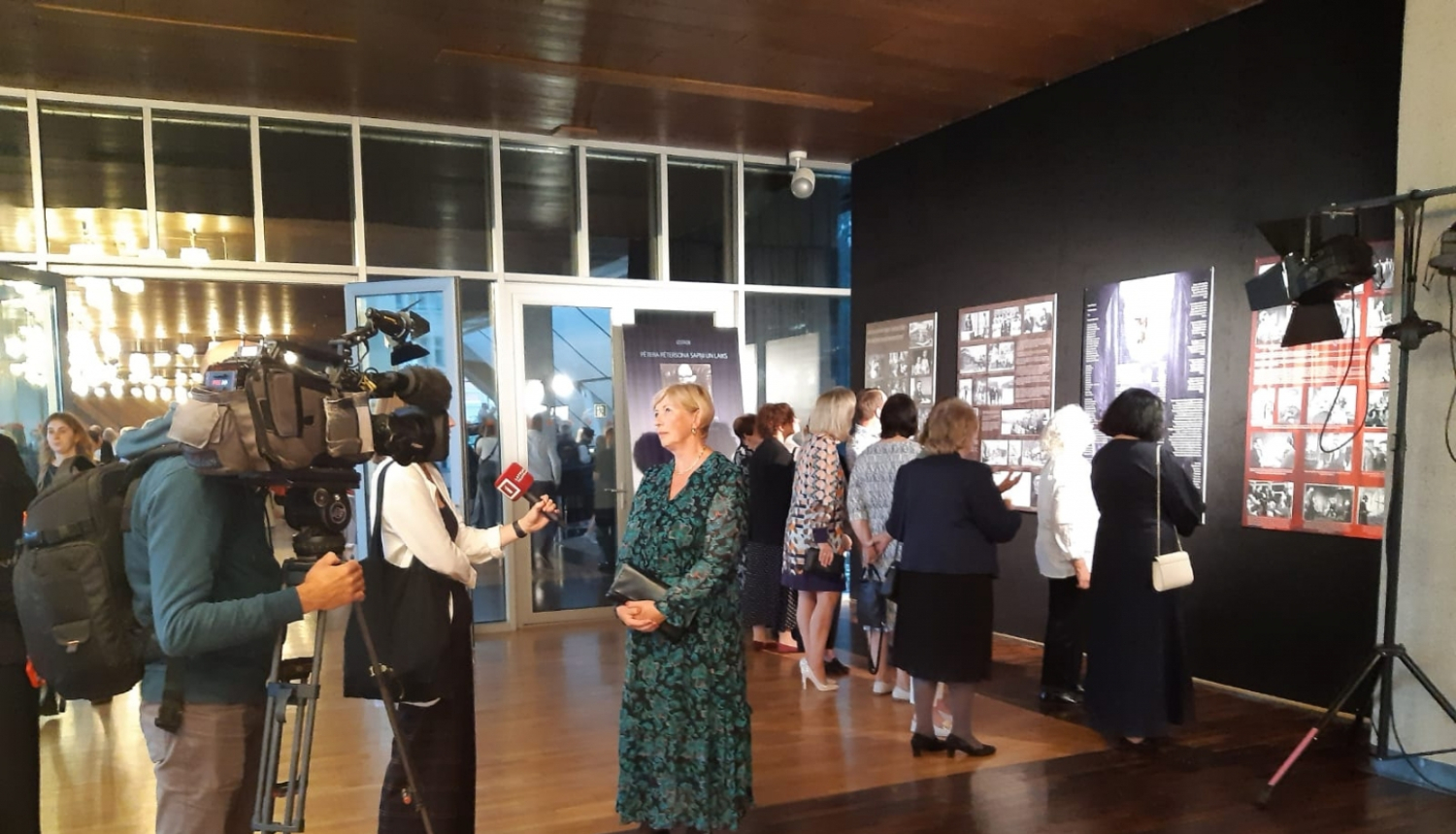 Krāsaina bilde, Pētera Pētersona izstādes atklāšana Dailes teātrī, K.Pētersone sniedz interviju žurnalistiem, fonā izstādes stendi un apmeklētāji