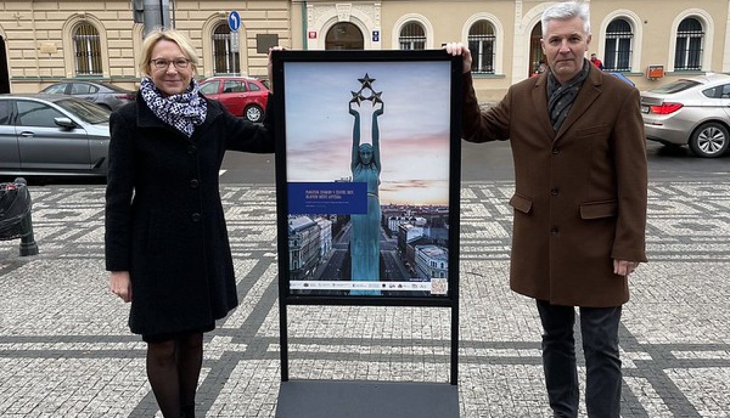 Sievietis un vīrietis pie plakāta ar brīvības pieminekļa attēlu